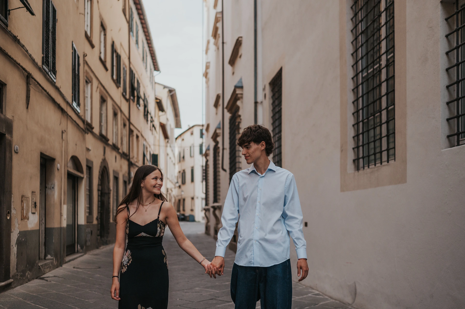 Alice and Pablo smile at one another as they walk hand in hand past the old town’s shuttered buildings. From Alice and Pablo’s Lucca anniversary photoshoot.