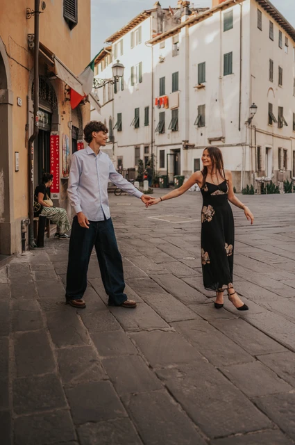 Lucca couple photoshoot around an old-town square Alice and Pablo hold hands at the corner of a Lucca square surrounded by pale buildings and shopfronts. From Alice and Pablo’s Lucca anniversary photoshoot.