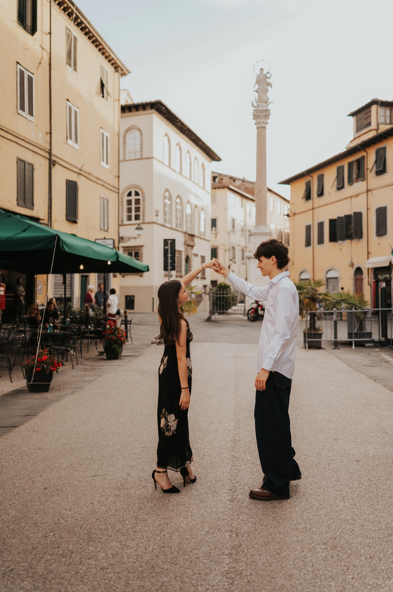 Pablo lifts Alice’s hand as she turns in a Lucca square, with a tall stone column behind them. From Alice and Pablo’s Lucca anniversary photoshoot.