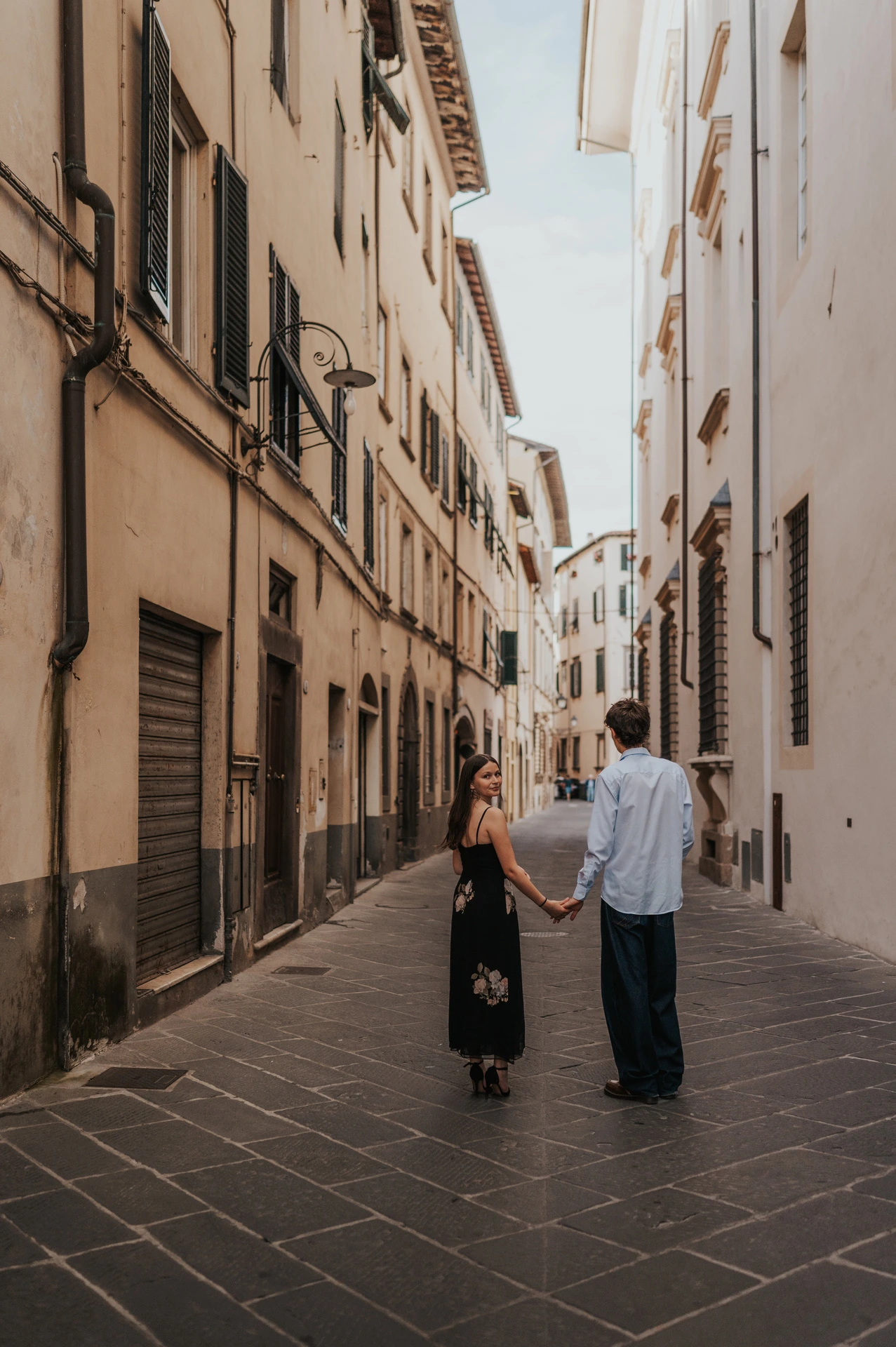 Alice and Pablo walk away hand in hand down a narrow Lucca lane lined with pale buildings. From Alice and Pablo’s Lucca anniversary photoshoot.
