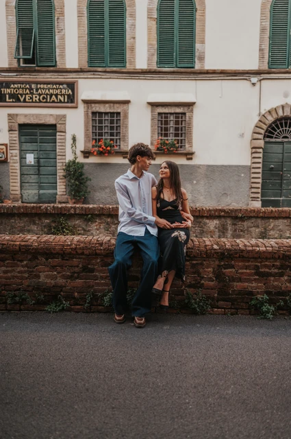 Lucca couple photoshoot beside Via del Fosso’s brick wall Pablo stands beside Alice as she sits on a low brick wall along Via del Fosso. From Alice and Pablo’s Lucca anniversary photoshoot.