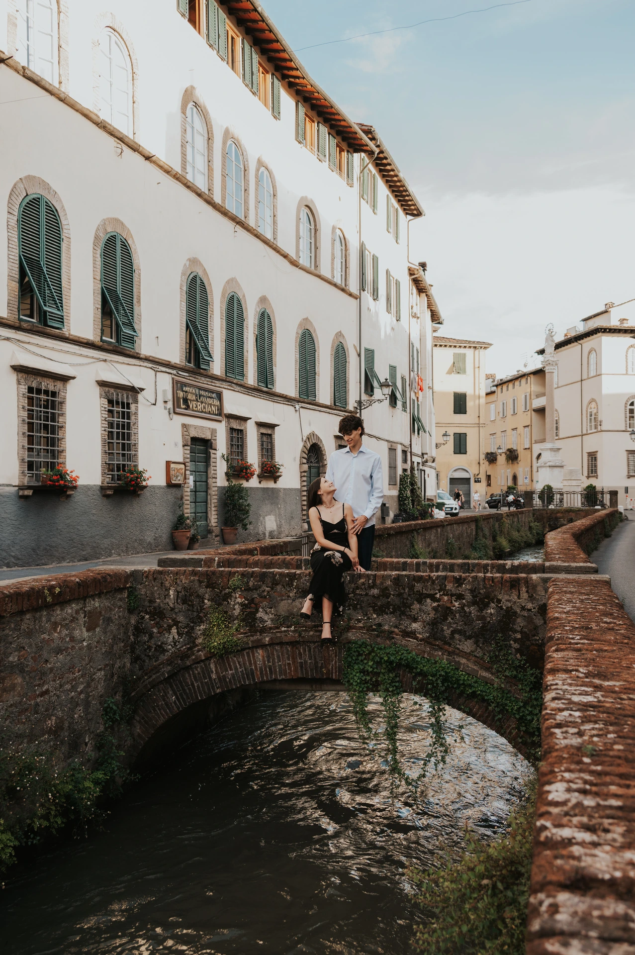 Alice and Pablo pose on a small brick bridge above the canal on Via del Fosso, beside green-shuttered buildings. From Alice and Pablo’s Lucca anniversary photoshoot.