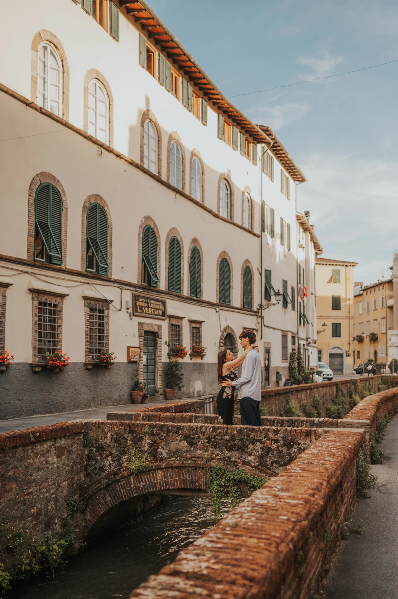 Lucca’s narrow canal passes beneath a brick bridge beside white buildings with green shutters. From Alice and Pablo’s Lucca anniversary photoshoot.