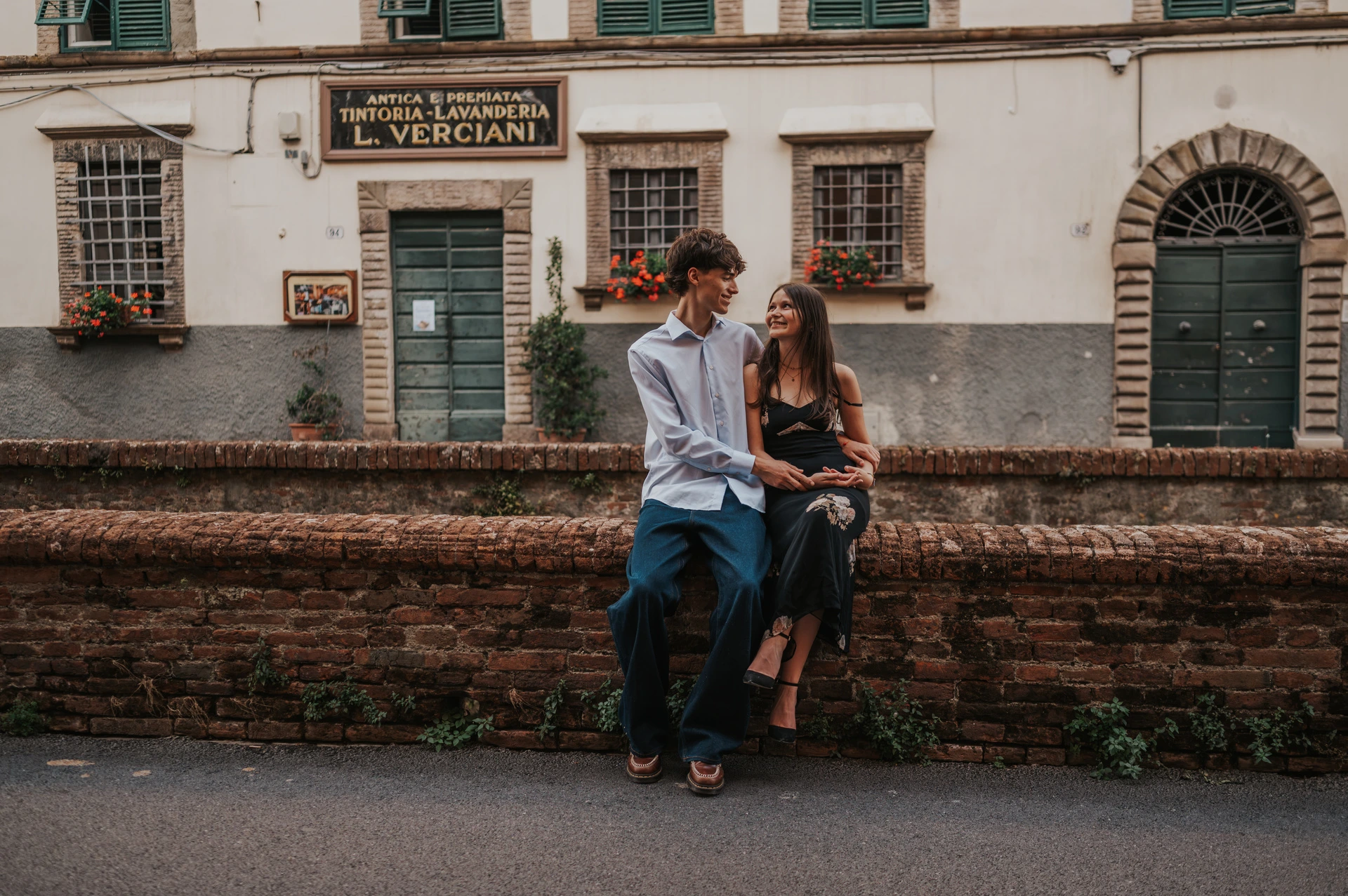 Pablo leans towards Alice as she sits on a brick wall beneath flower boxes and green shutters. From Alice and Pablo’s Lucca anniversary photoshoot.