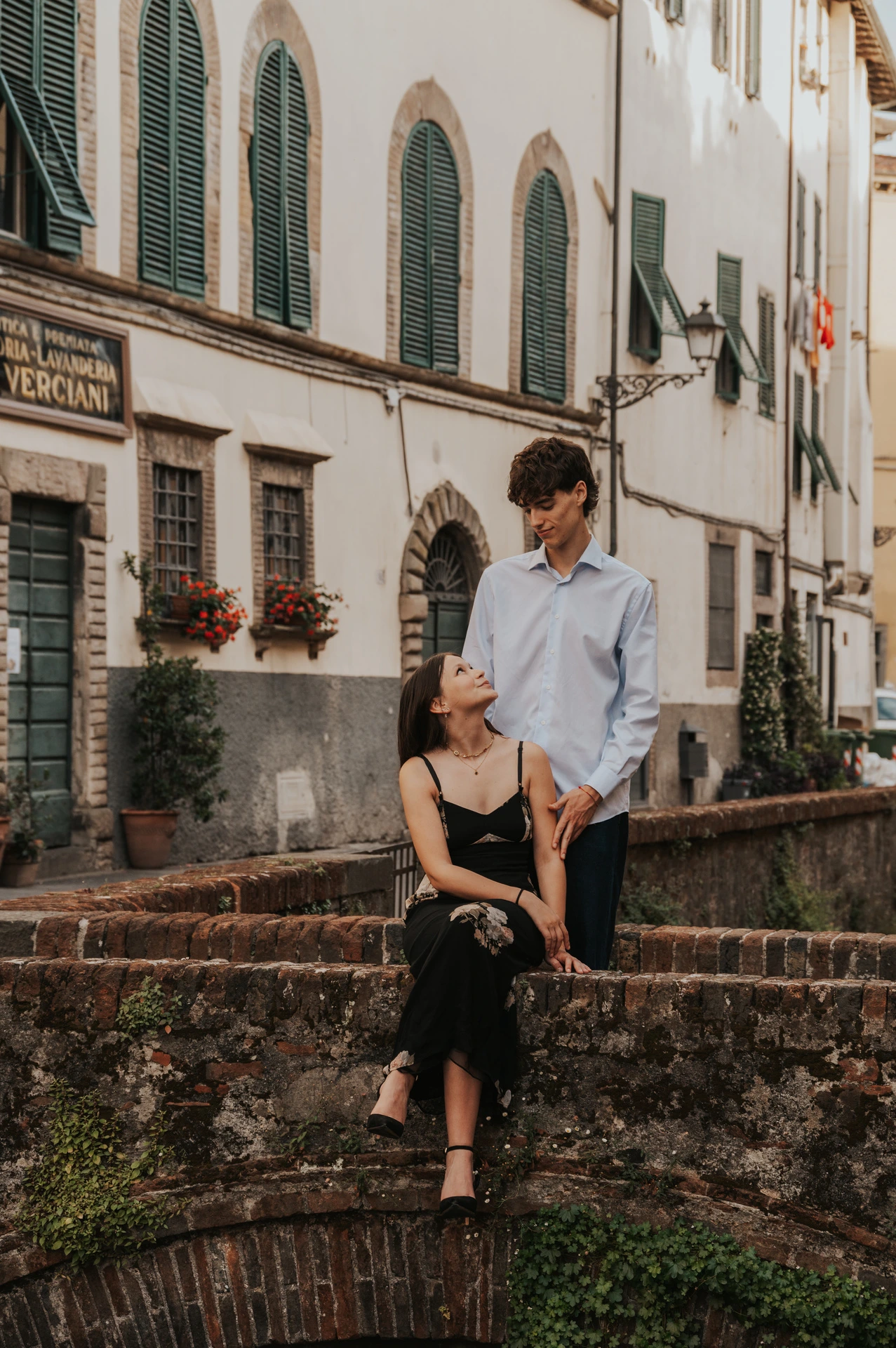 Alice sits on the brick bridge beside Pablo, with green shutters and flower boxes behind them. From Alice and Pablo’s Lucca anniversary photoshoot.