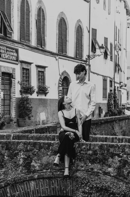 Lucca couple photoshoot on a canal bridge, in black and white Black-and-white portrait of Alice seated beside Pablo on the brick bridge along Via del Fosso. From Alice and Pablo’s Lucca anniversary photoshoot.