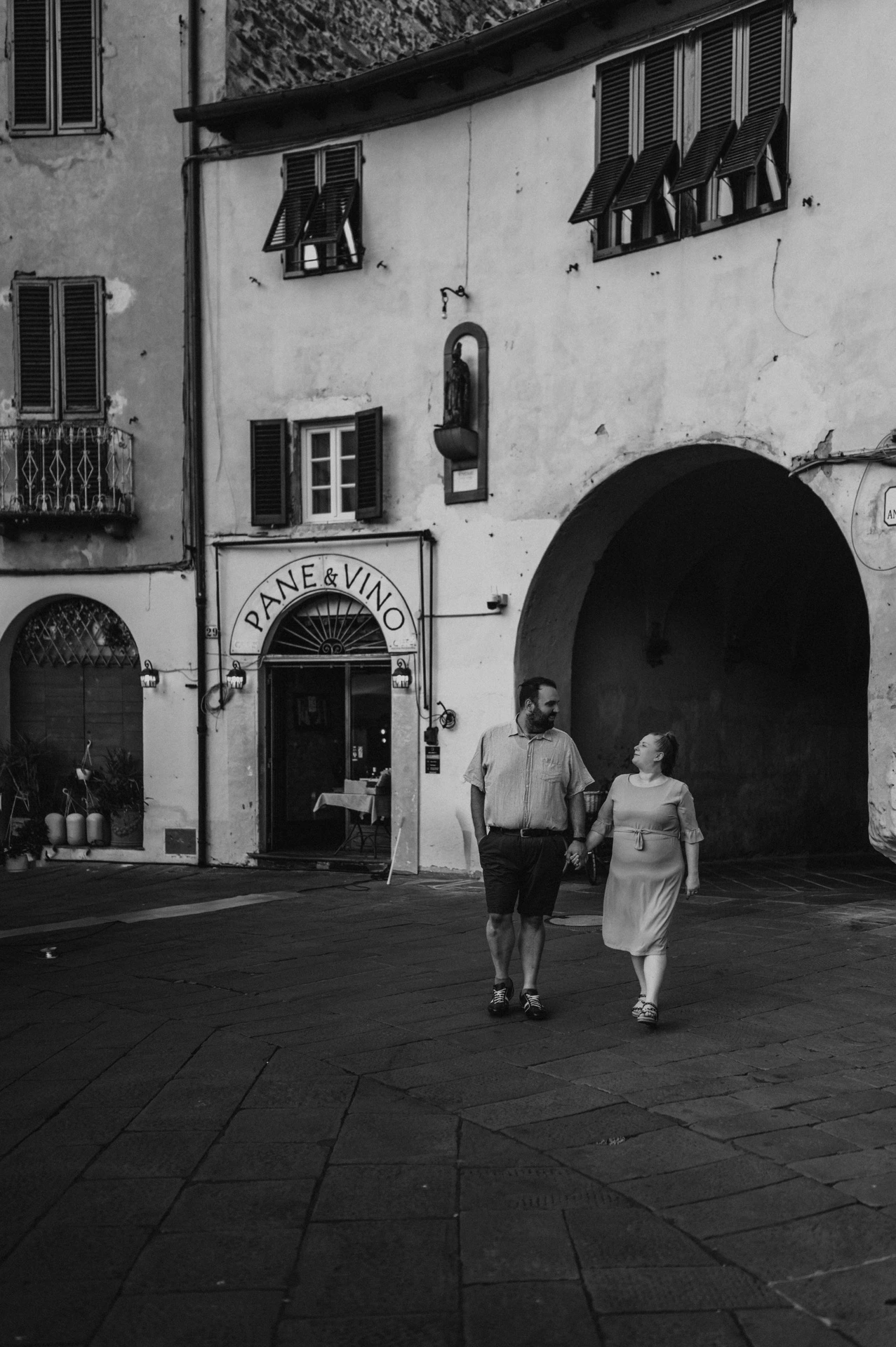 Black-and-white photograph of the couple walking beside the shaded arch and shuttered façades of Piazza dell’Anfiteatro during a couple photoshoot in Lucca.
