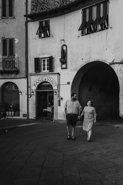 Lucca couple photoshoot: a shaded arch in Piazza dell’Anfiteatro in black and white Black-and-white photograph of the couple walking beside the shaded arch and shuttered façades of Piazza dell’Anfiteatro during a couple photoshoot in Lucca.