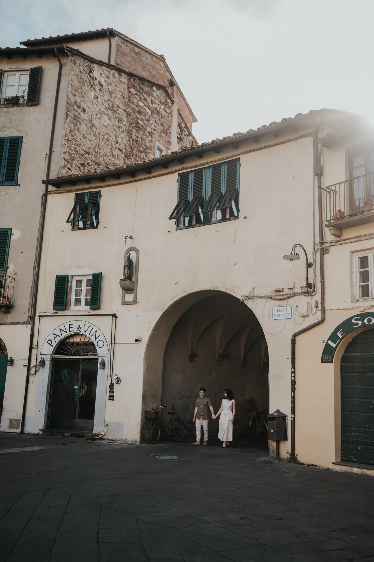 A wide view of the couple beneath the tall façades beside an arched entrance to Piazza dell’Anfiteatro during a couple photoshoot in Lucca.