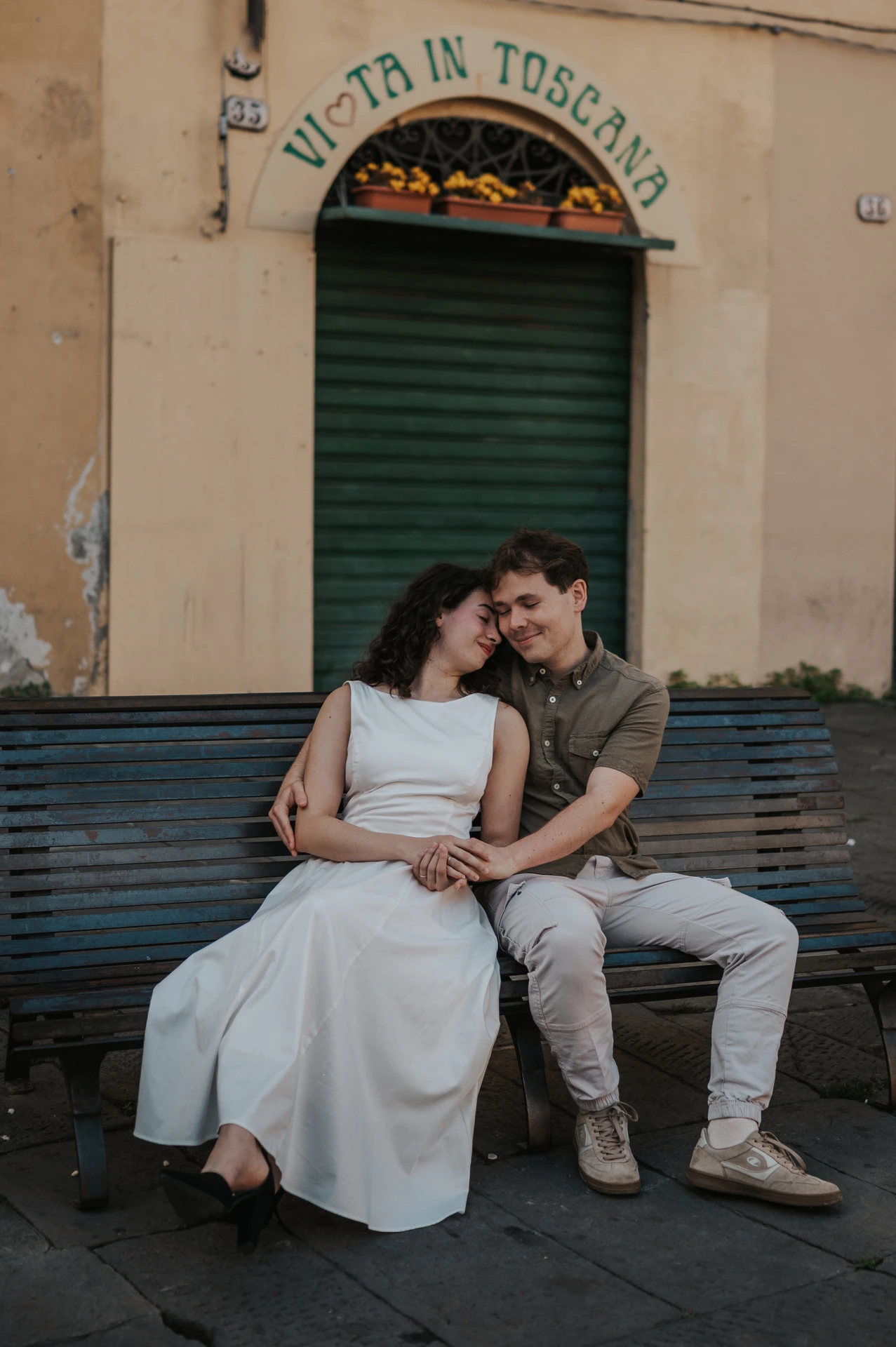 The couple leaning close on a green bench beside Piazza dell’Anfiteatro, with their hands resting together during a couple photoshoot in Lucca.