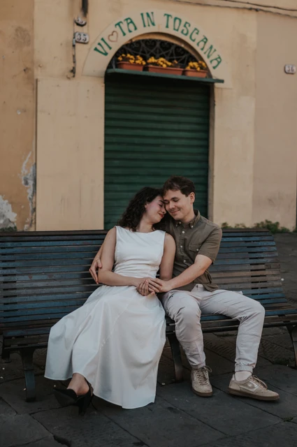The couple leaning close on a green bench beside Piazza dell’Anfiteatro, with their hands resting together during a couple photoshoot in Lucca.