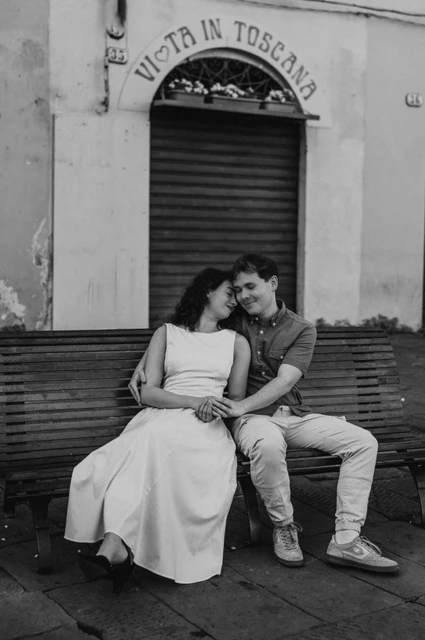 Lucca couple photoshoot: a bench portrait beside the piazza in black and white Black-and-white photograph of the couple sitting close on a bench in front of a shuttered doorway beside Piazza dell’Anfiteatro during a couple photoshoot in Lucca.