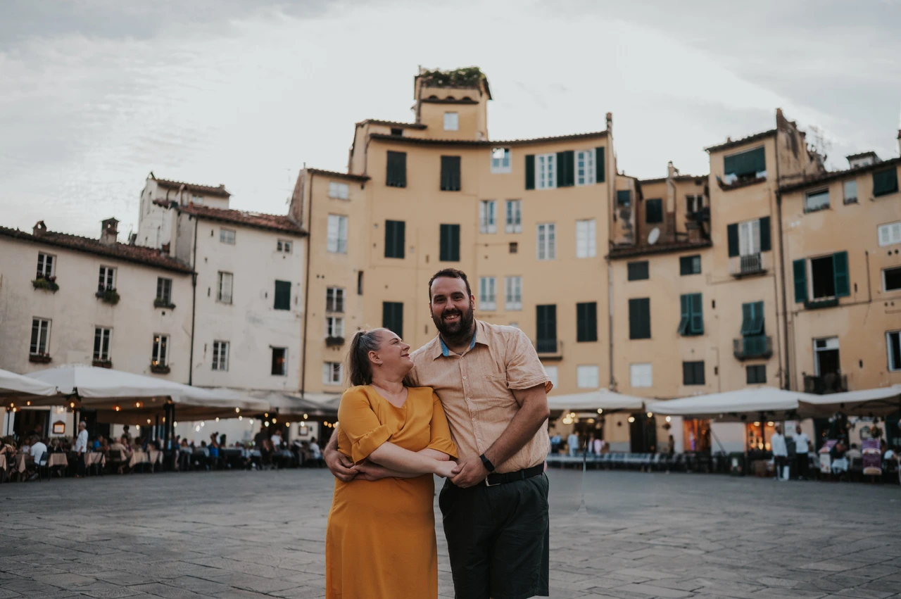 Lucca couple photoshoot: a couple portrait in Piazza dell’Anfiteatro The couple standing close together in Piazza dell’Anfiteatro, with the surrounding façades and cafés behind them during a couple photoshoot in Lucca.