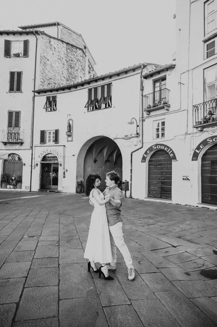 Lucca couple photoshoot: a portrait with the piazza’s arch behind in black and white Black-and-white photograph of the couple standing close together in Piazza dell’Anfiteatro with an arch and shuttered buildings behind them during a couple photoshoot in Lucca.