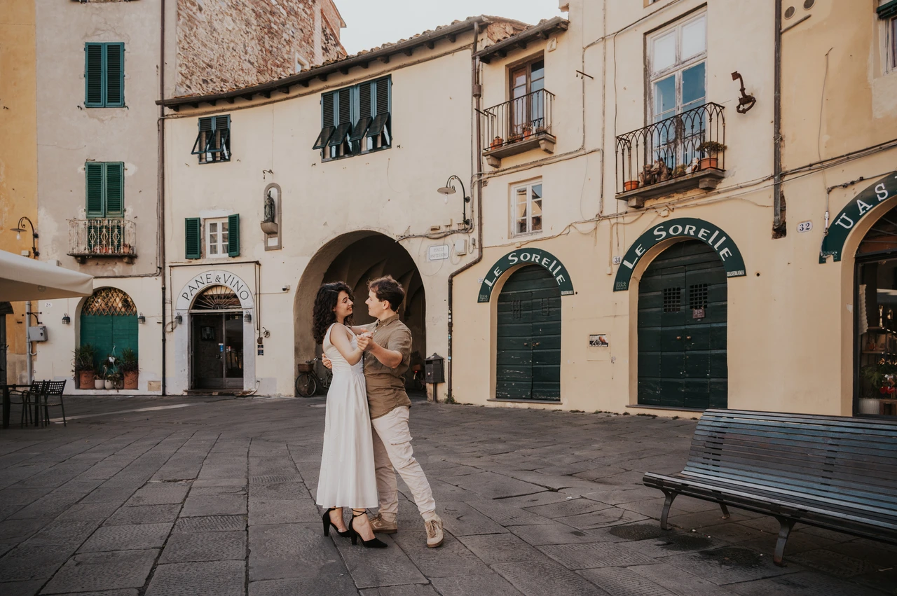 Lucca couple photoshoot: a wide portrait beside the piazza arch A wide portrait of the couple embracing beside Piazza dell’Anfiteatro’s arched entrance and a bench during a couple photoshoot in Lucca.