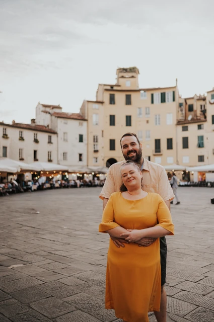 The man standing behind his partner with his arms around her in Piazza dell’Anfiteatro during a couple photoshoot in Lucca.