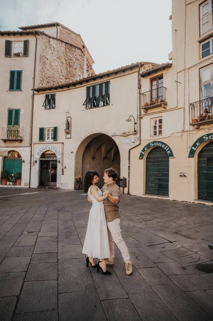 Lucca couple photoshoot: an embrace in Piazza dell’Anfiteatro The couple embracing in Piazza dell’Anfiteatro with pale façades and an arched entrance behind them during a couple photoshoot in Lucca.