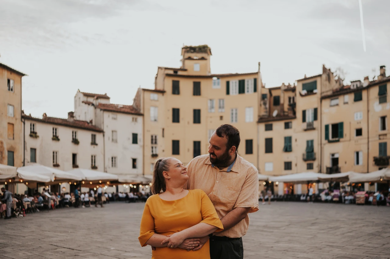 Lucca couple photoshoot: an embrace with the piazza’s façades behind A wide portrait of the couple looking towards each other in Piazza dell’Anfiteatro, with his arms around her during a couple photoshoot in Lucca.