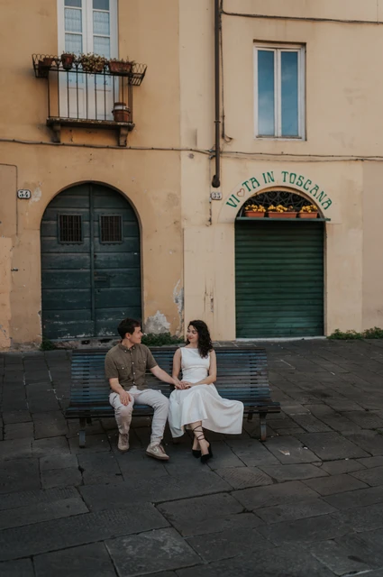Lucca couple photoshoot: a green bench beside Piazza dell’Anfiteatro The couple sitting together on a green bench below shuttered doors beside Piazza dell’Anfiteatro during a couple photoshoot in Lucca.