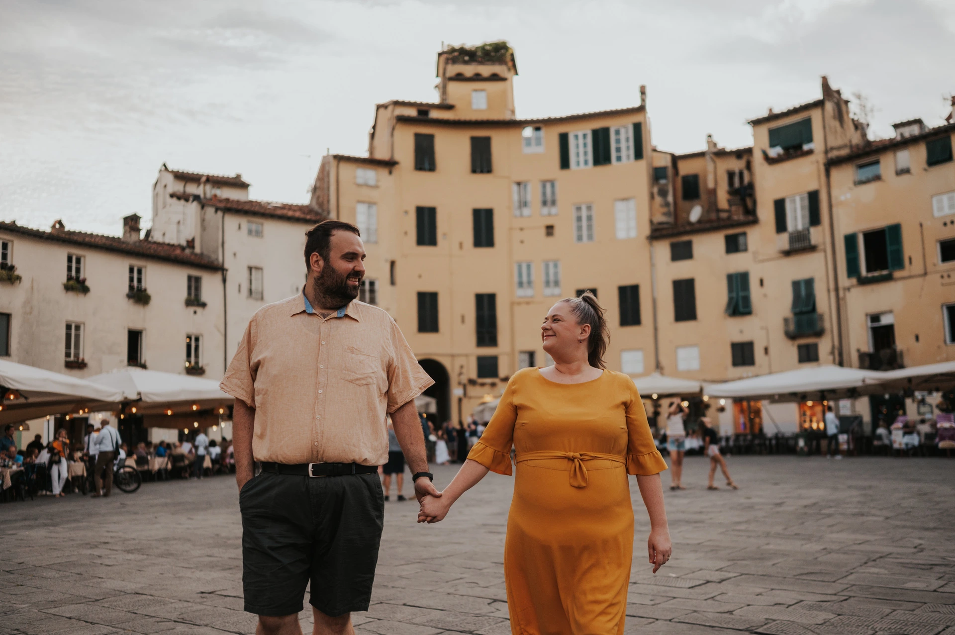 Lucca couple photoshoot: a shared look in Piazza dell’Anfiteatro The couple looking towards each other while walking hand in hand in Piazza dell’Anfiteatro during a couple photoshoot in Lucca.