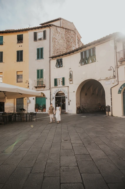 Lucca couple photoshoot: walking across the open piazza A wide view of the couple walking across the open paving with Piazza dell’Anfiteatro’s pale buildings around them during a couple photoshoot in Lucca.