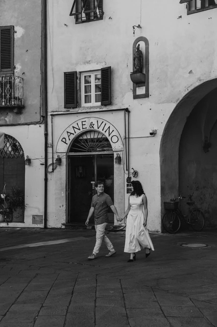 Lucca couple photoshoot: a walk beside the piazza’s shopfronts in black and white Black-and-white photograph of the couple walking hand in hand beside shuttered windows and a wine-shop doorway in Piazza dell’Anfiteatro during a couple photoshoot in Lucca.