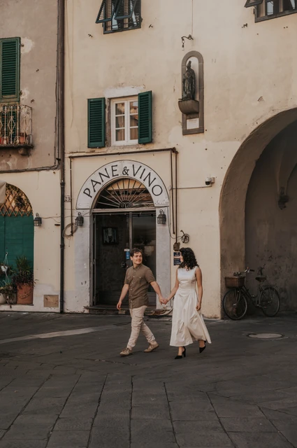Lucca couple photoshoot: a walk past the piazza’s shuttered shops The couple walking hand in hand past shuttered shops and pale façades in Piazza dell’Anfiteatro during a couple photoshoot in Lucca.