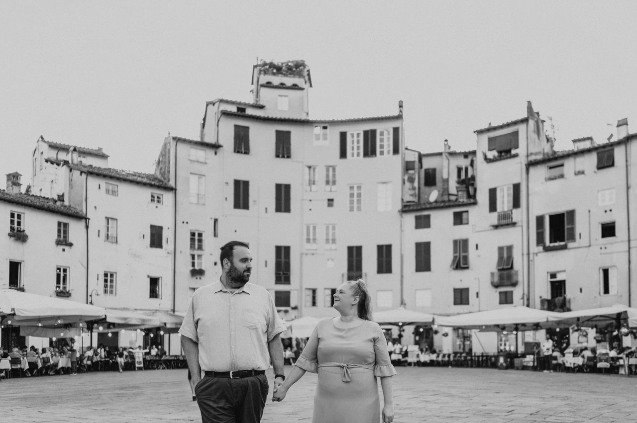 Lucca couple photoshoot: walking together in Piazza dell’Anfiteatro in black and white Black-and-white photograph of the couple walking hand in hand with Piazza dell’Anfiteatro’s curved row of façades behind them during a couple photoshoot in Lucca.