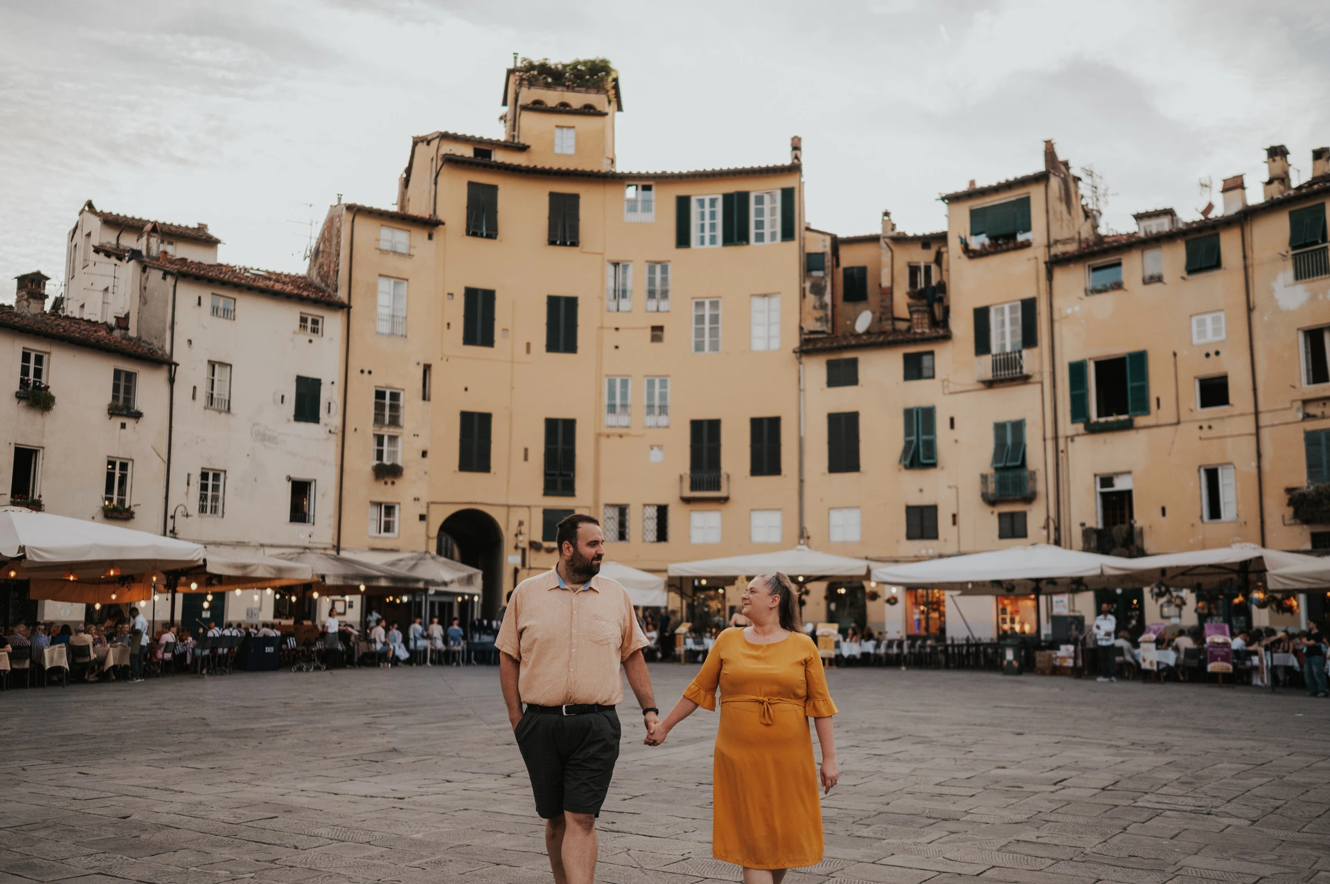 The couple walking hand in hand across Piazza dell’Anfiteatro, with yellow and cream façades behind them during a couple photoshoot in Lucca.