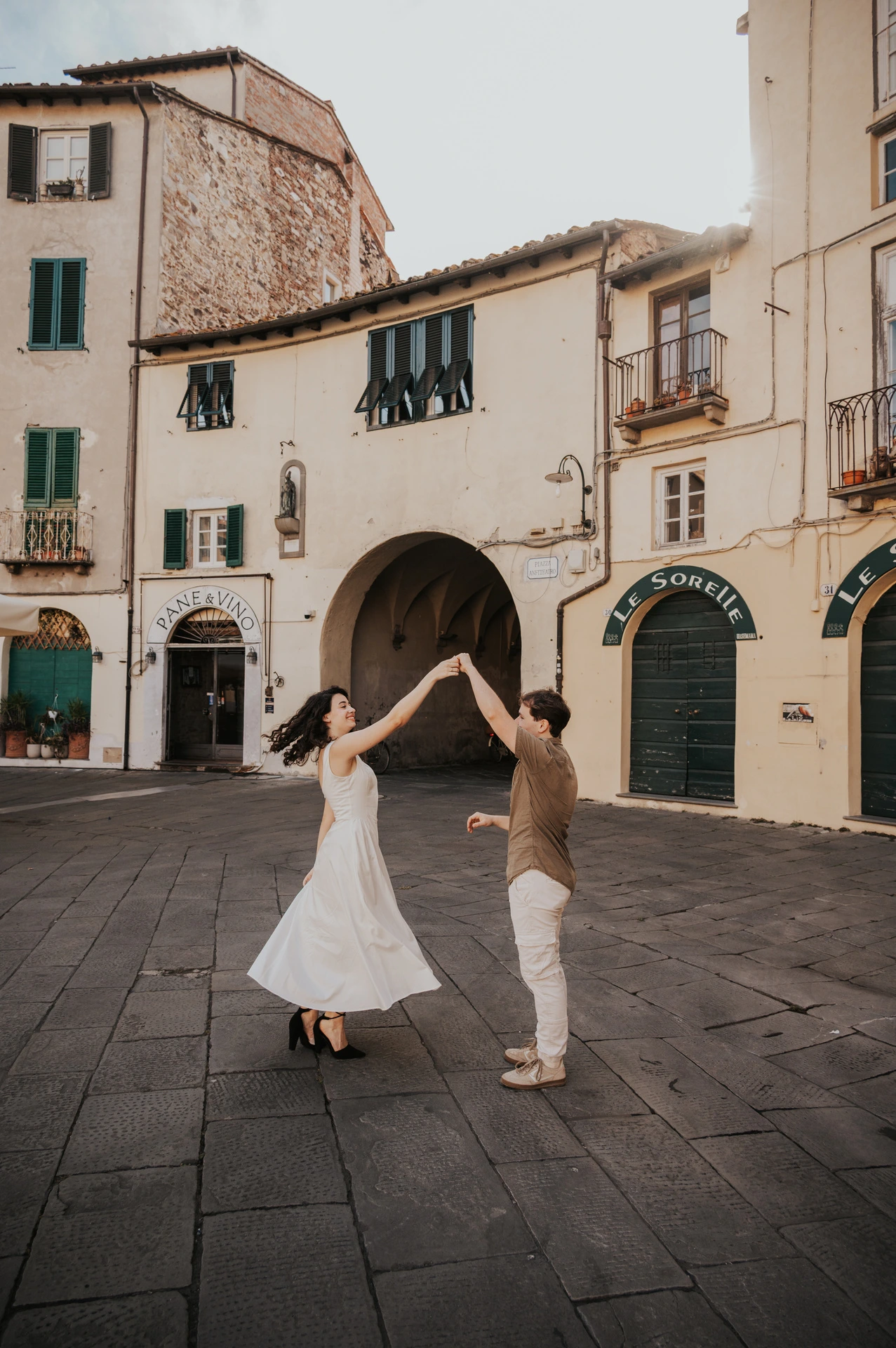 The woman’s white dress sweeping out as her partner raises her hand in Piazza dell’Anfiteatro during a couple photoshoot in Lucca.