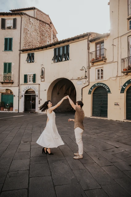 Lucca couple photoshoot: a white dress turning in the piazza The woman’s white dress sweeping out as her partner raises her hand in Piazza dell’Anfiteatro during a couple photoshoot in Lucca.