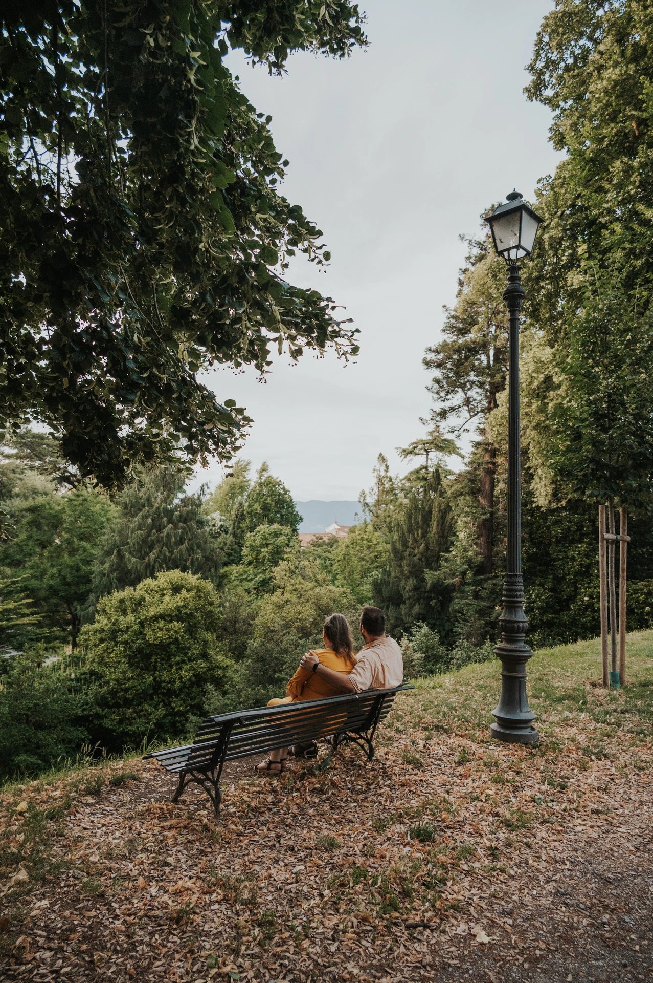 The couple sitting close on a bench beneath leafy trees beside a path on Lucca’s city walls during a couple photoshoot in Lucca.