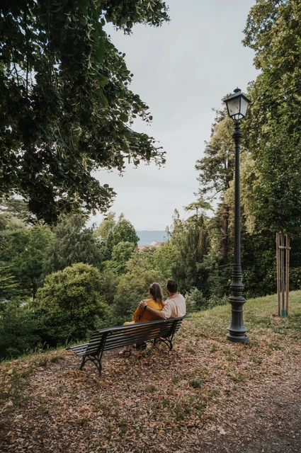 The couple sitting close on a bench beneath leafy trees beside a path on Lucca’s city walls during a couple photoshoot in Lucca.