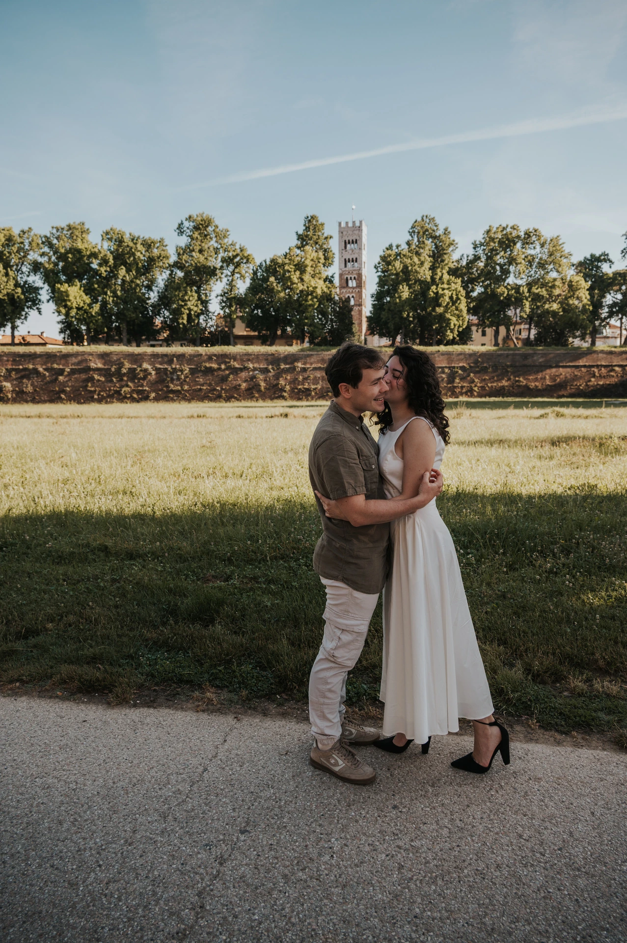 A full-length portrait of the couple standing close beside the grass near the city walls during a couple photoshoot in Lucca.