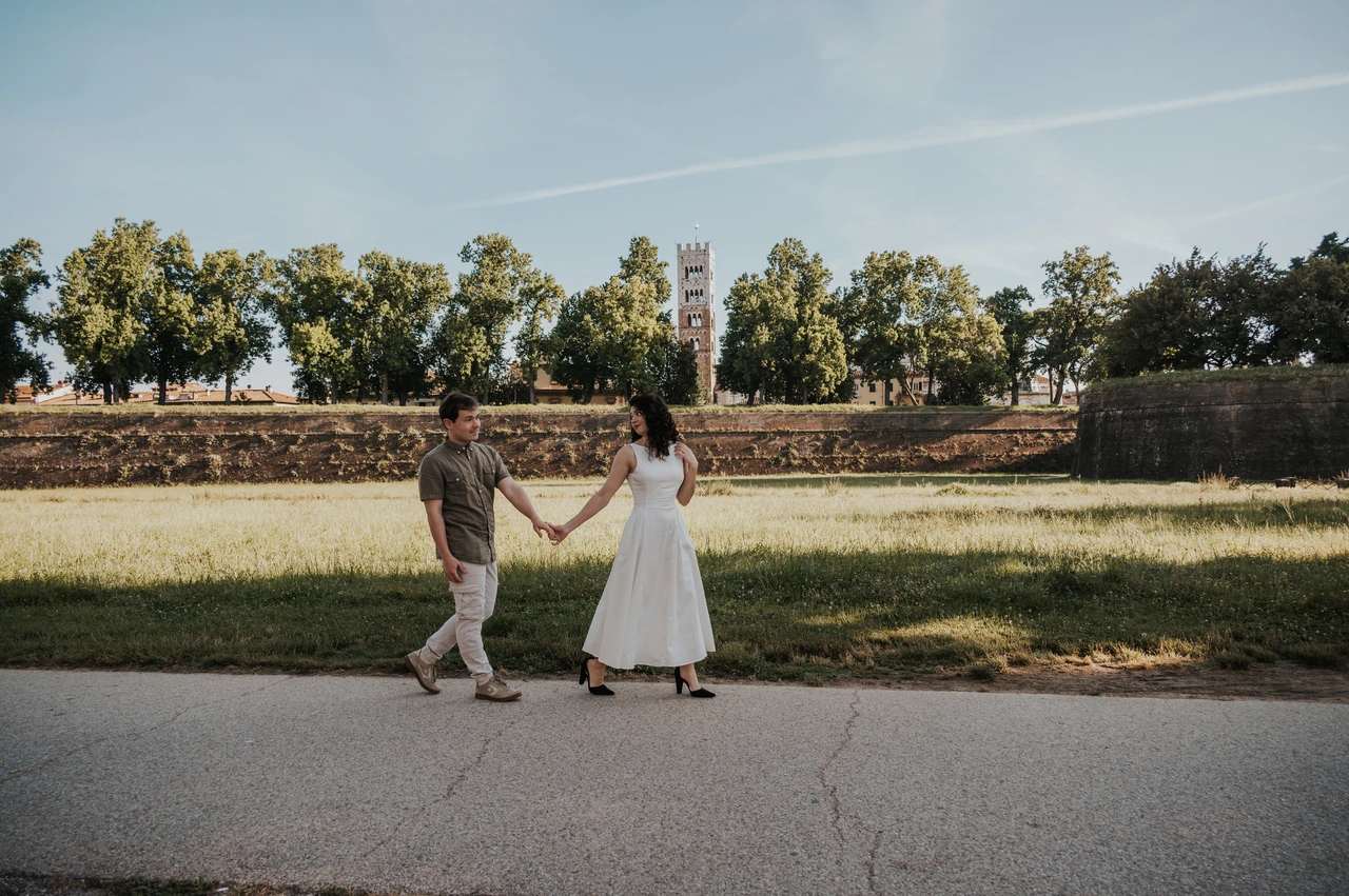 Lucca couple photoshoot: a wide portrait beside the green city walls The couple standing apart and holding hands on a path beside the green city walls during a couple photoshoot in Lucca.