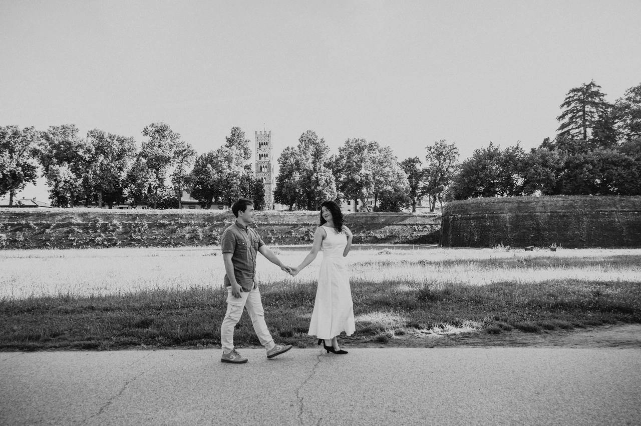 Lucca couple photoshoot: a landscape portrait on the city-wall path in black and white Black-and-white photograph of a wide view of the couple walking together beside the trees and grass of the city walls during a couple photoshoot in Lucca.