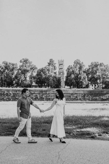 Lucca couple photoshoot: a walk beside the city walls in black and white Black-and-white photograph of the couple walking hand in hand beside the grass and trees near the city walls during a couple photoshoot in Lucca.