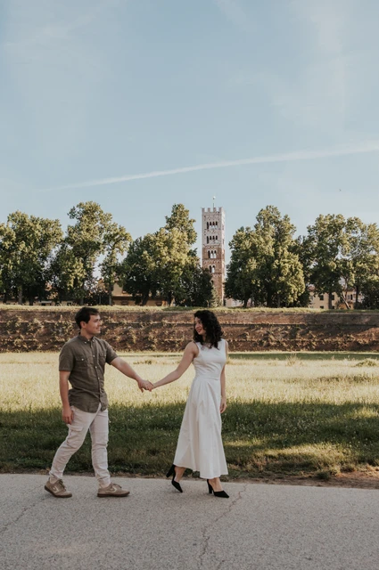 Lucca couple photoshoot: holding hands with San Frediano’s tower behind The couple holding hands on a path with grass, trees and San Frediano’s bell tower behind them during a couple photoshoot in Lucca.