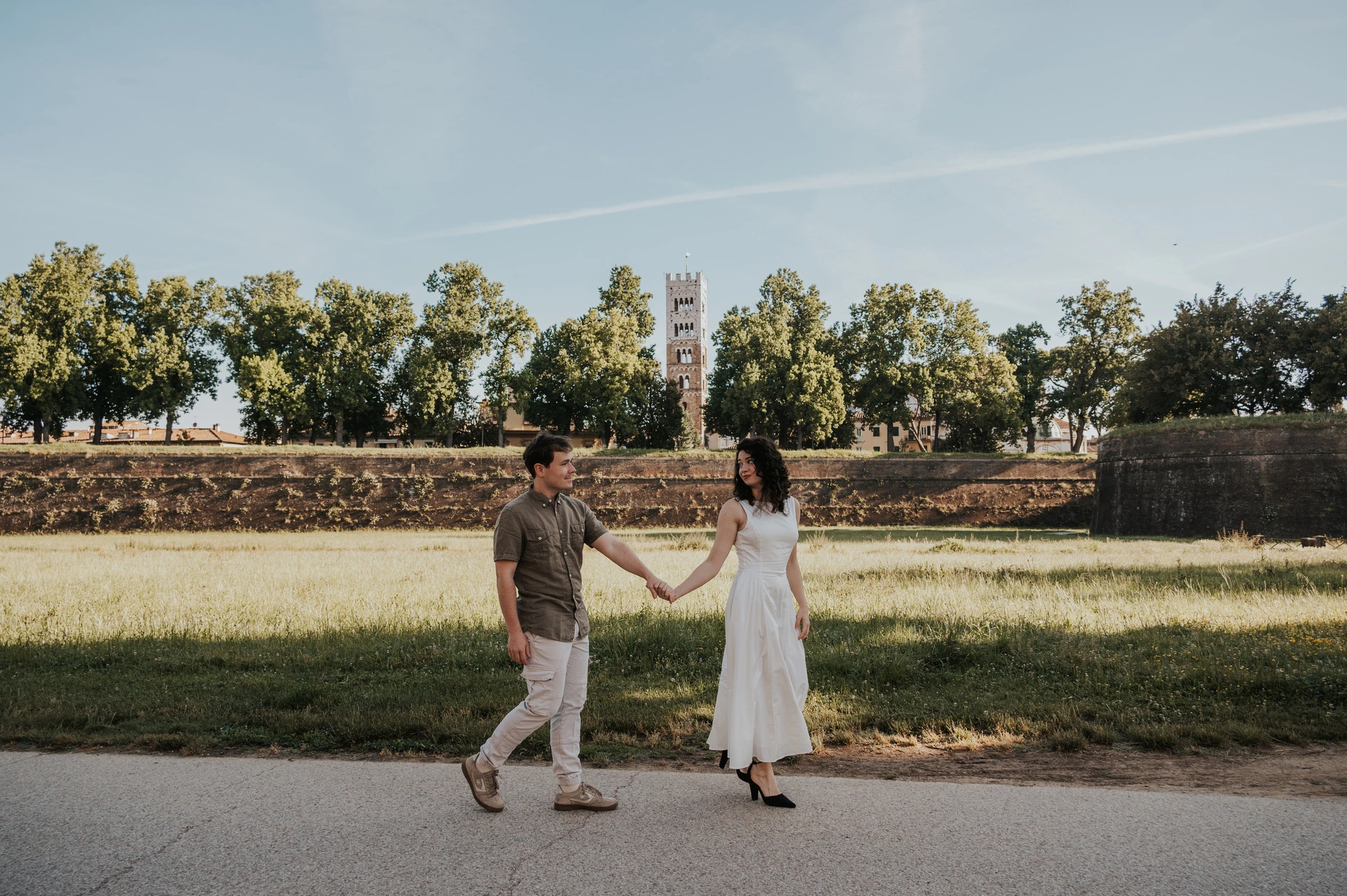 Lucca couple photoshoot: a wide view of the path by the walls A wide view of the couple walking together beside the grass with San Frediano’s tower in the distance during a couple photoshoot in Lucca.