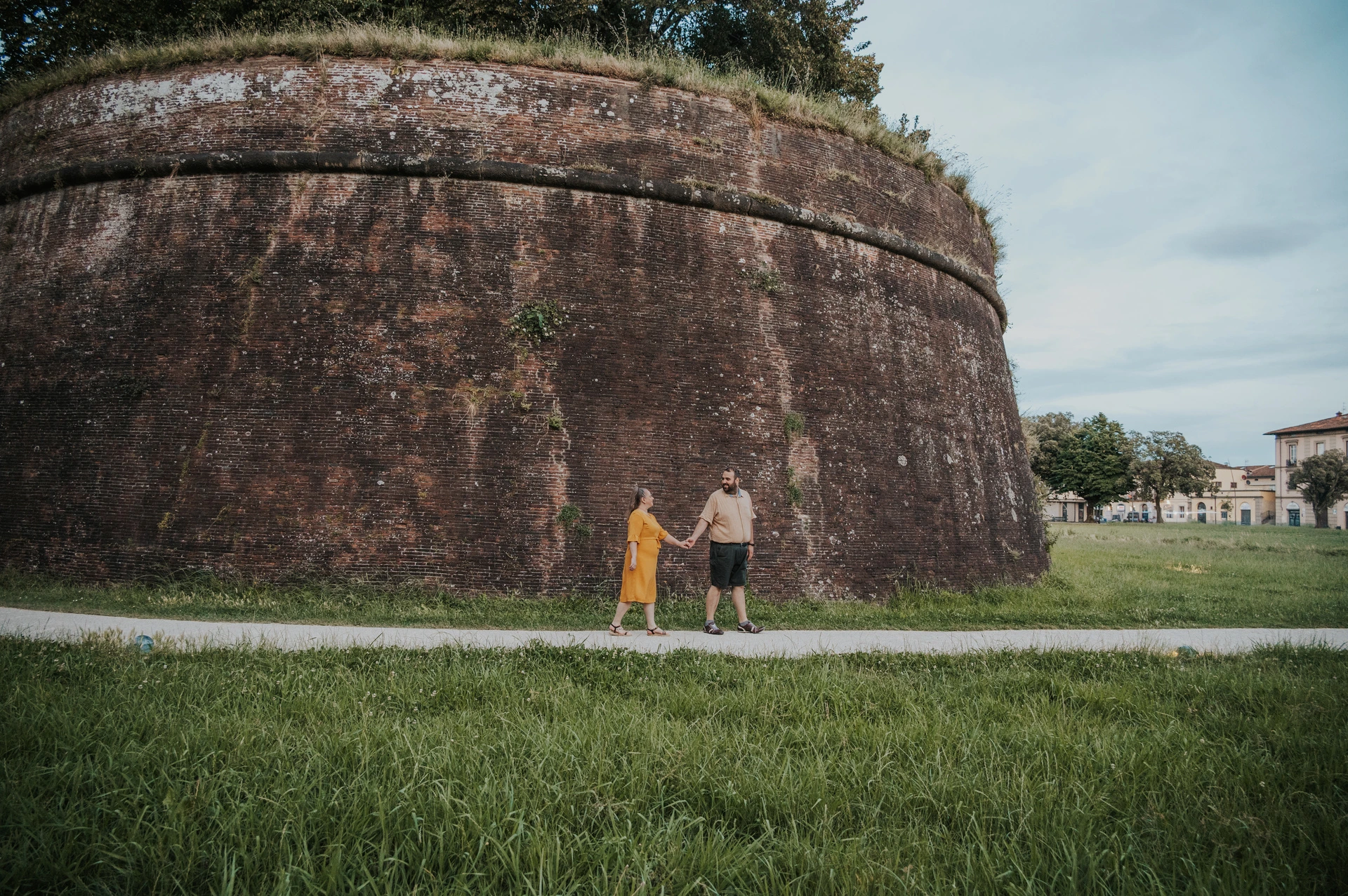 Lucca couple photoshoot: the curved city walls and green grass A wide view of the couple holding hands beside Lucca’s curved brick walls, with grass and trees around them during a couple photoshoot in Lucca.