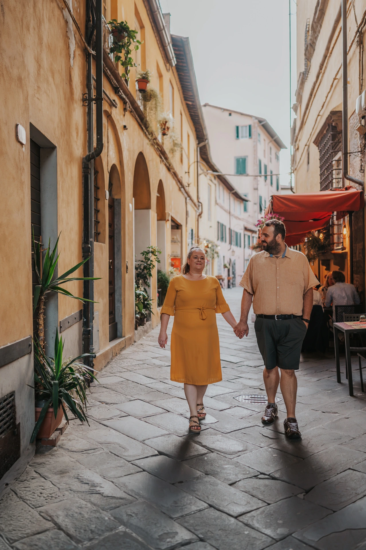 The couple walking towards the camera beneath an arch between warm-coloured buildings in the old town during a couple photoshoot in Lucca.