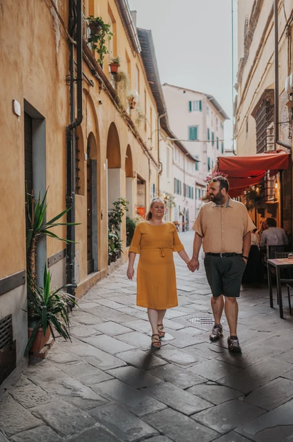 Lucca couple photoshoot: walking beneath an old-town archway The couple walking towards the camera beneath an arch between warm-coloured buildings in the old town during a couple photoshoot in Lucca.
