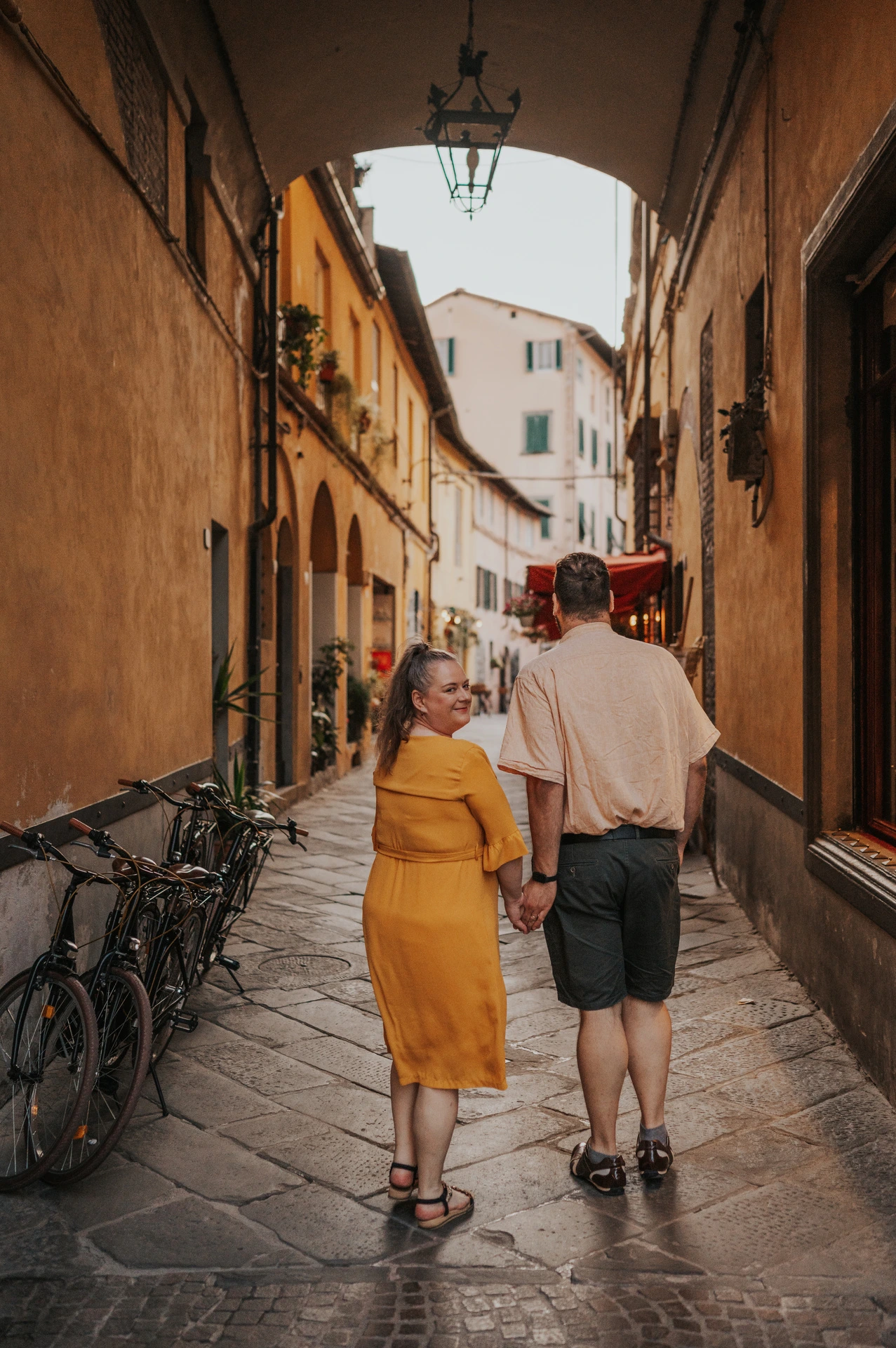 A full-length view of the couple walking away hand in hand through an arched lane in the old town during a couple photoshoot in Lucca.