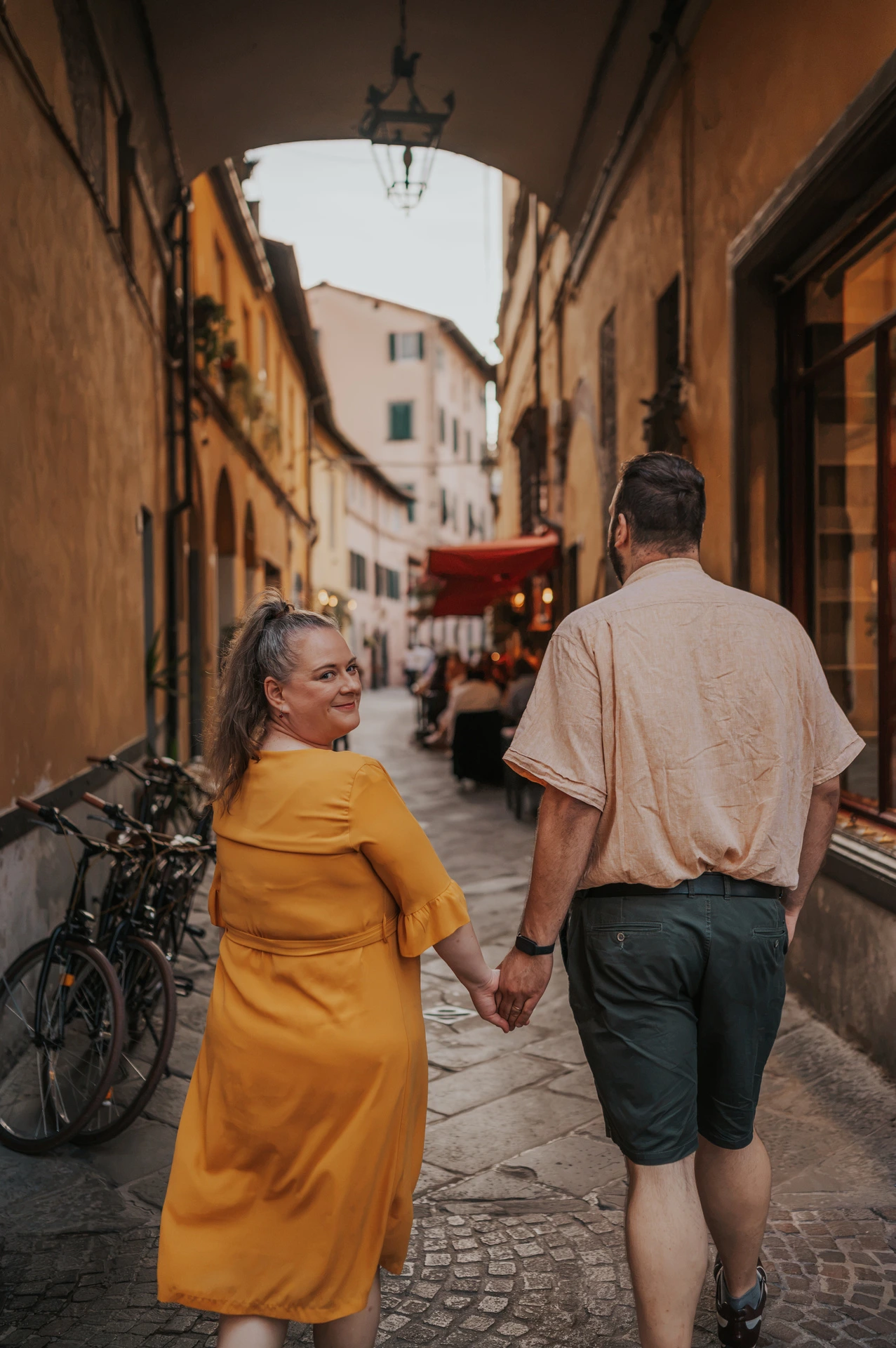 The couple walking hand in hand through an arched old-town lane, with the woman looking back during a couple photoshoot in Lucca.