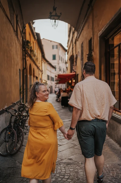 Lucca couple photoshoot: looking back beneath an old-town arch The couple walking hand in hand through an arched old-town lane, with the woman looking back during a couple photoshoot in Lucca.