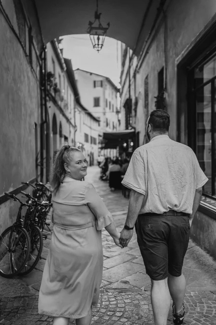 Black-and-white photograph of the couple walking hand in hand beneath an arch in a narrow old-town street lined with bicycles during a couple photoshoot in Lucca.