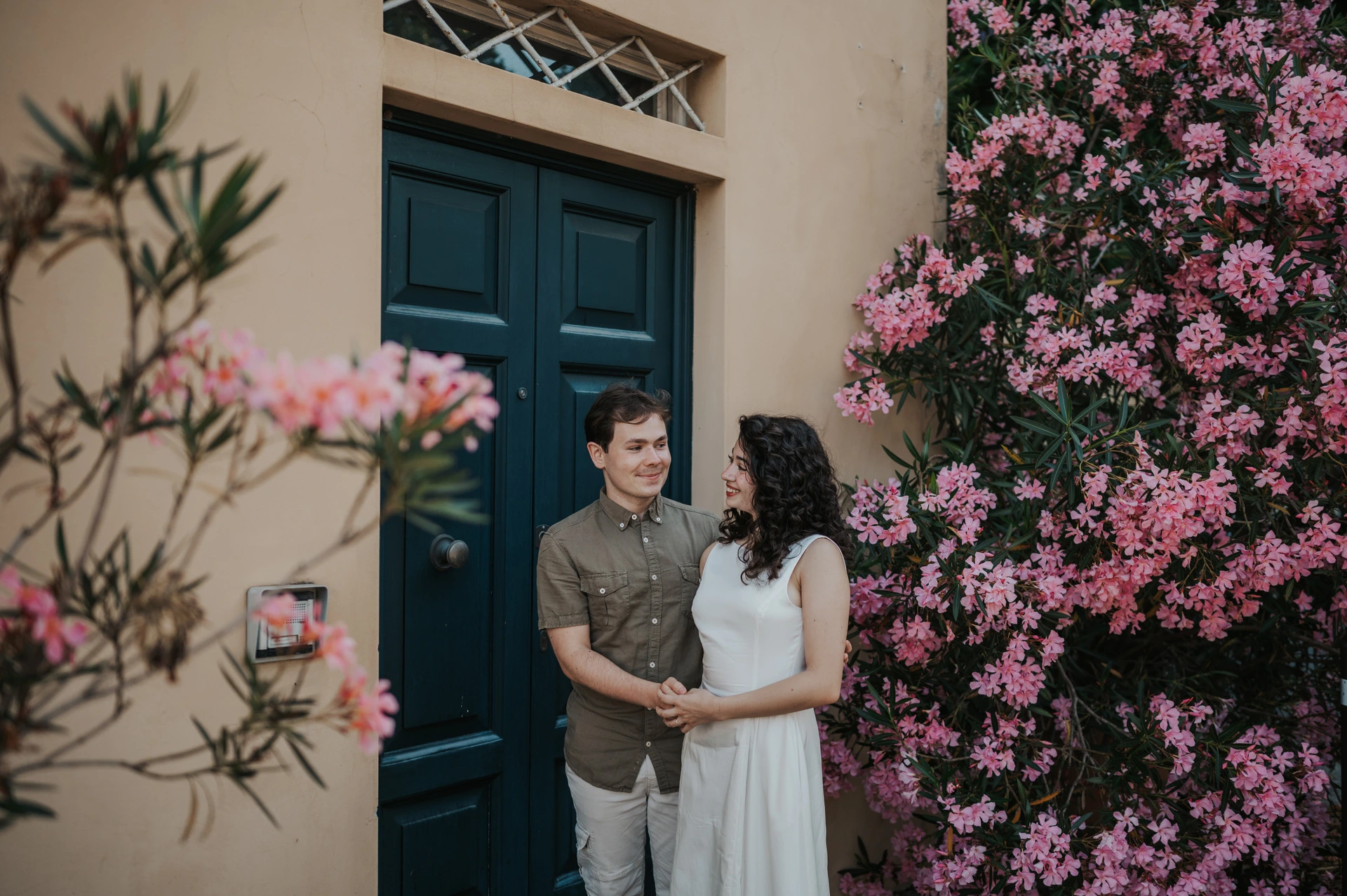 A wide portrait of the couple standing close beside a green door framed by pink flowers during a couple photoshoot in Lucca.