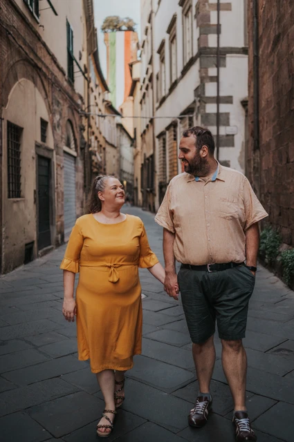 Lucca couple photoshoot: a close portrait in an old-town street The couple looking towards each other as they walk hand in hand along a narrow street, with Guinigi Tower in the distance during a couple photoshoot in Lucca.
