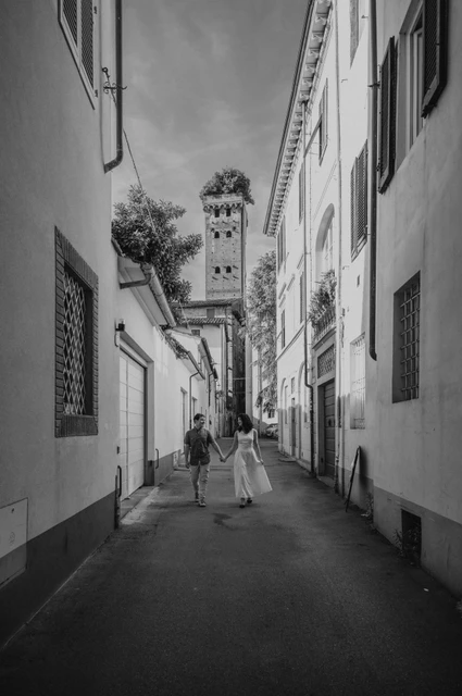 Black-and-white photograph of the couple walking along a narrow street lined with pale buildings, with Guinigi Tower beyond during a couple photoshoot in Lucca.