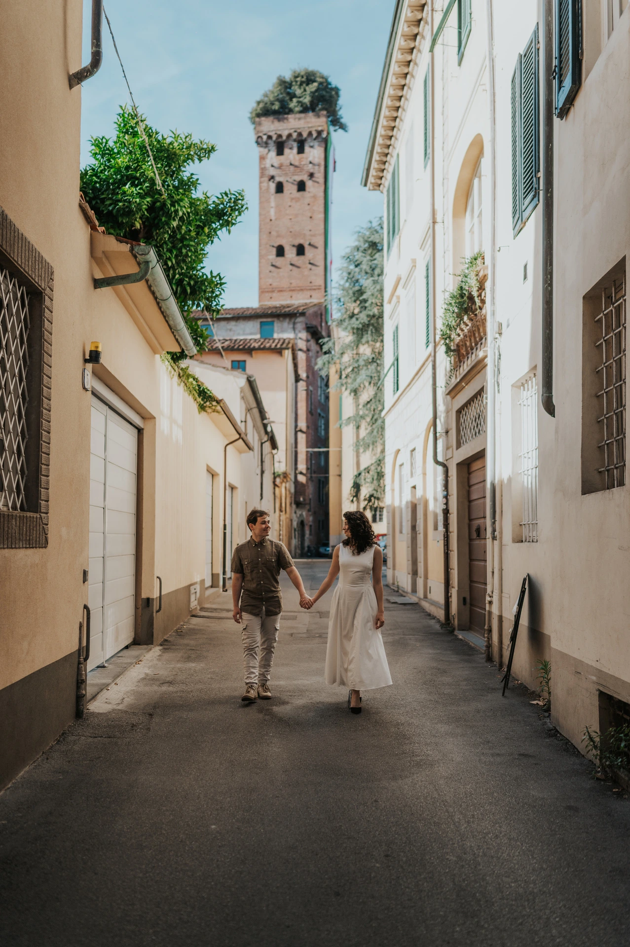 The couple walking along a narrow old-town street with Guinigi Tower and its rooftop trees behind them during a couple photoshoot in Lucca.